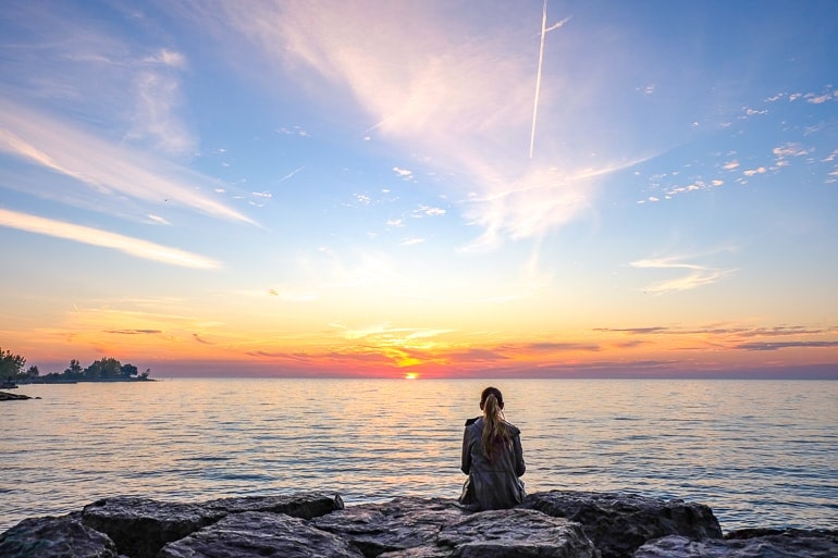 girl sitting on rocks with sunrise behind over lake ontario.