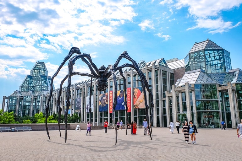 large metal spider in front of glass building with blue sky above.