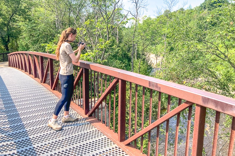 woman with camera and hiking shoes standing on metal bridge.