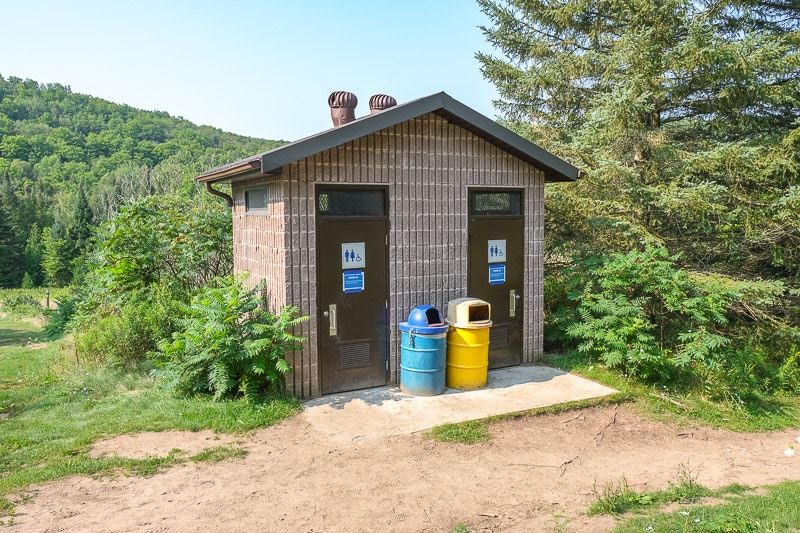 small bathroom building with garbage cans and green forest behind.