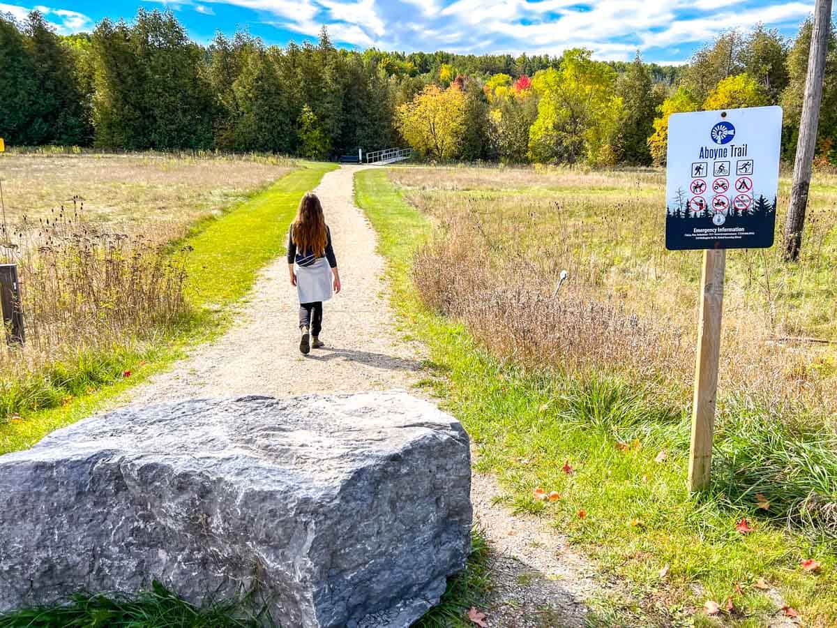 woman on hiking path through green field with large stone and trail sign in foreground.